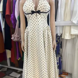 Vintage-inspired polka dot halter-neck maxi dress with black polka dots, a plunging neckline, and a contrasting black bow or ribbon accent at the bust.