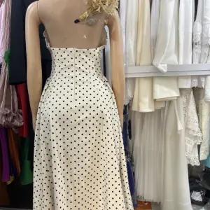 Vintage-inspired polka dot halter-neck maxi dress with black polka dots, a plunging neckline, and a contrasting black bow or ribbon accent at the bust.